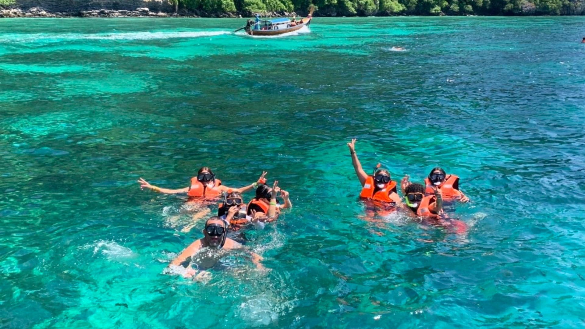 Siete bañistas con gafas de buceo, seis con chaleco naranja, saludan en el agua turquesa ante una barca de cola larga